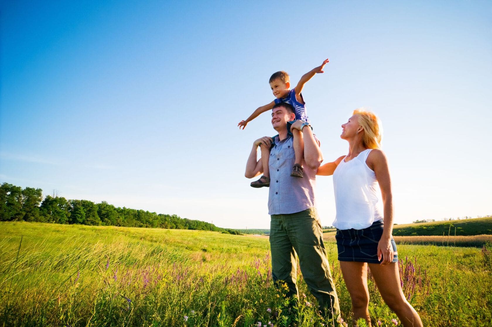 Glad familie står på grøn mark med skov i baggrunden og kigger op mod en blå himmel.
